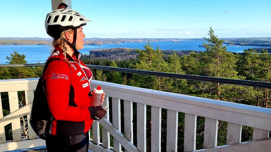 Ina Mäkinen seisoo korkealla näköalatornissa pyöräilykypärä päässä. Ympärillä levittäytyy kaunis, suomalainen järvimaisema.